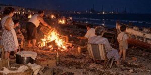 Vecinos de un bloque obrero alrededor de una hoguera de cajas de pescado en la playa; al fondo, chimeneas de una fábrica.
