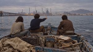 Tres jóvenes de espaldas en una barca de madera frente a la fábrica; uno de ellos estira el brazo con el pulgar levantado.