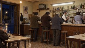 Interior de un bar de barrio: un camarero seca la barra, una mujer maneja la cafetera y tres hombres de espaldas toman el café; en la pared, un calendario y una televisión vieja.