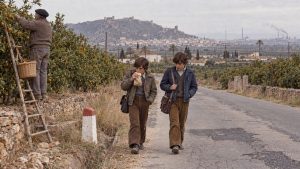 Dos chicos con la cartera vuelven andando por la carretera entre naranjos mientras un hombre coge naranjas de una escalera; al fondo, el castillo y las chimeneas.