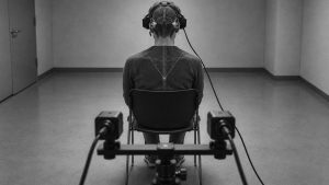 Persona sentada de espaldas con auriculares y el cerebro y la médula superpuestos, ante un sistema de dos cámaras sobre trípode