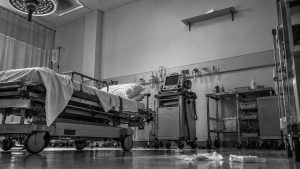 Habitación de hospital vacía vista desde el suelo: la cama deshecha, la lámpara quirúrgica encendida sobre ella, el carro del monitor y una estantería metálica al fondo